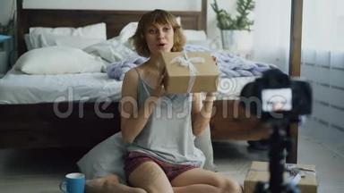 一位开朗的女士坐在床边，<strong>录制</strong>关于用dslr<strong>相机</strong>包装礼品盒的视频博客